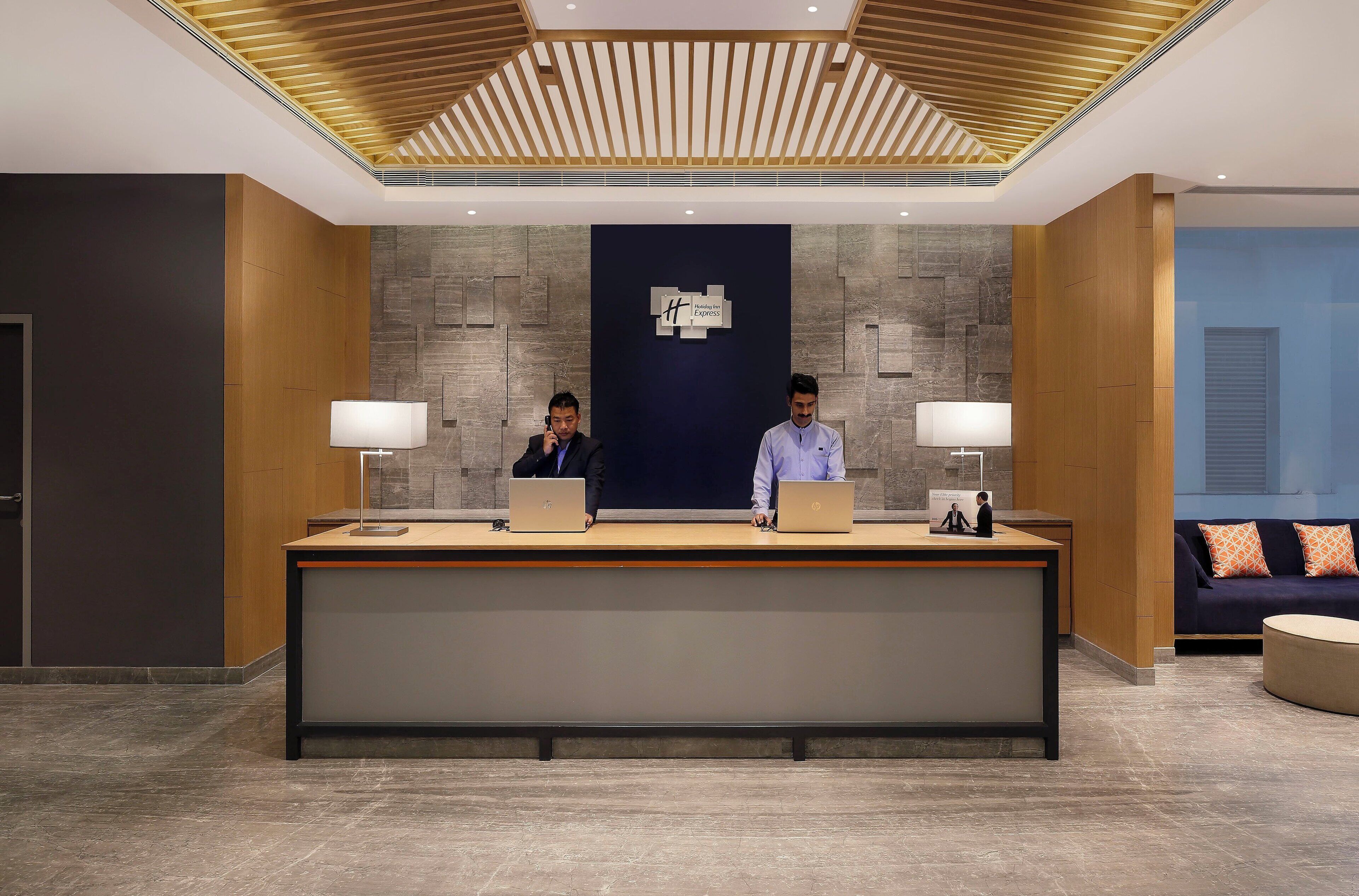 En hotellreception med två anställda, en modern takdesign och en väggbeklädnad av sten.