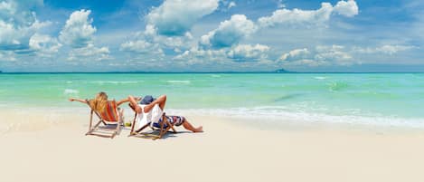 Escena de playa con dos personas relajándose en sillas de playa, cielo despejado y mar tranquilo.