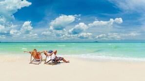Eine Strandansicht mit zwei Personen, die entspannt in Liegestühlen sitzen, einem klaren Himmel und ruhiger See.