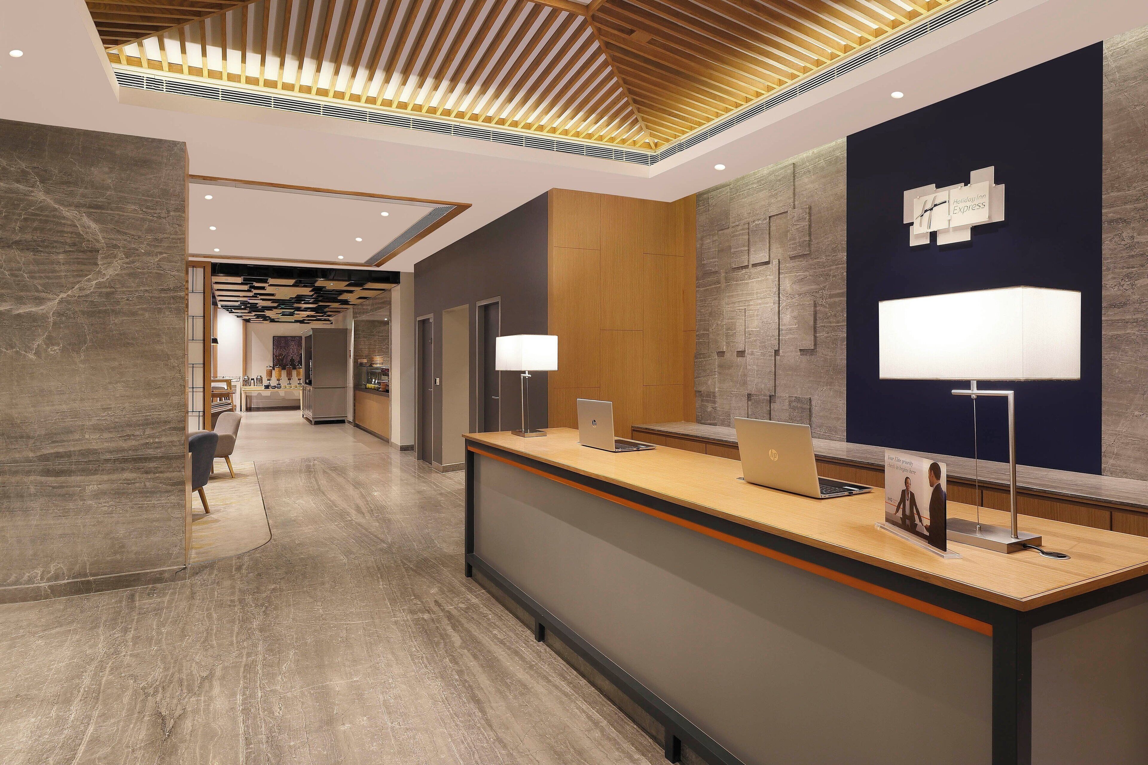 A modern hotel reception area with a wooden reception desk, two HP laptops, a wall clock, and a reception desk sign.
