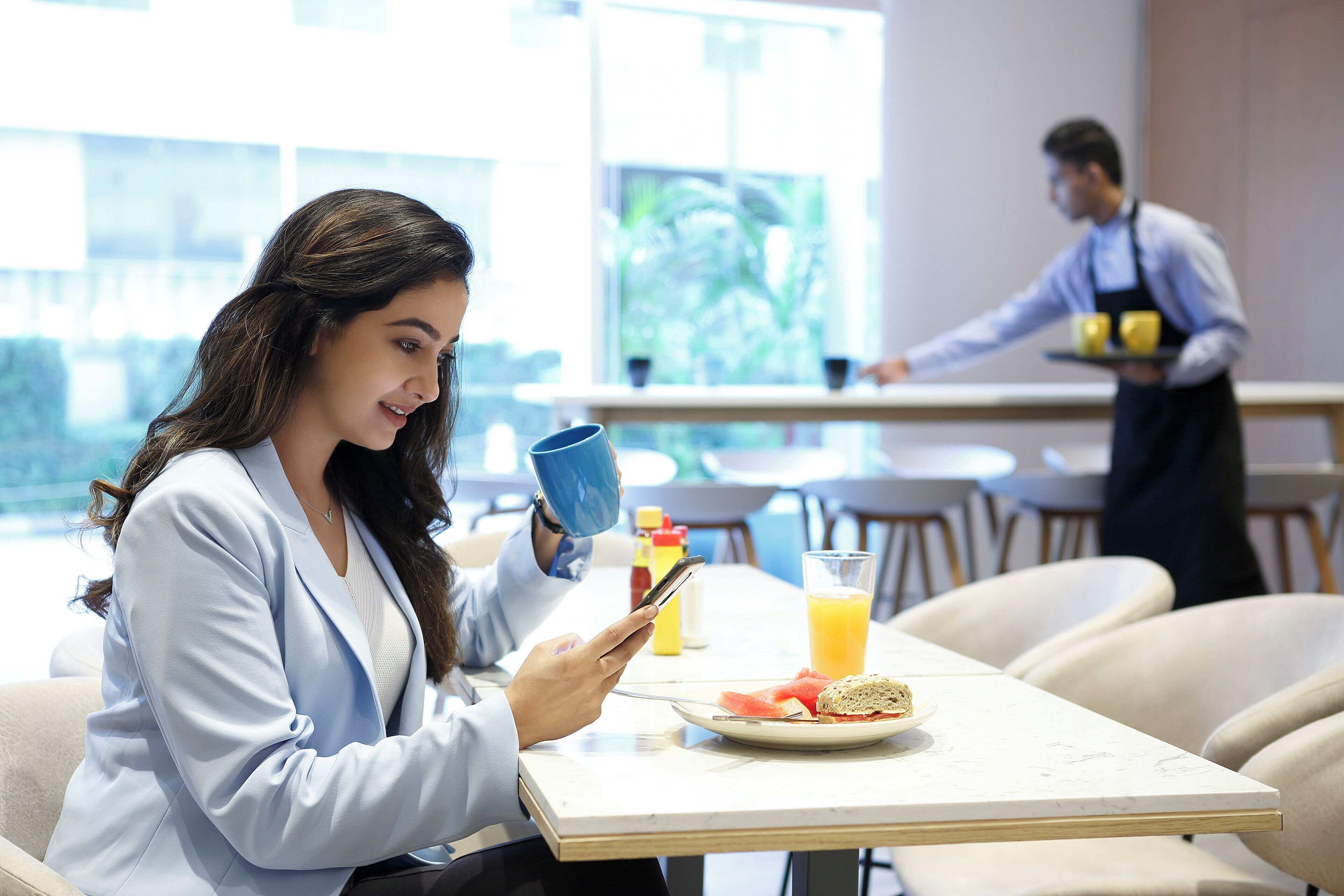 A person sitting at a table with a smartphone, a plate with food, and a drink. Another person is in the background near a counter.