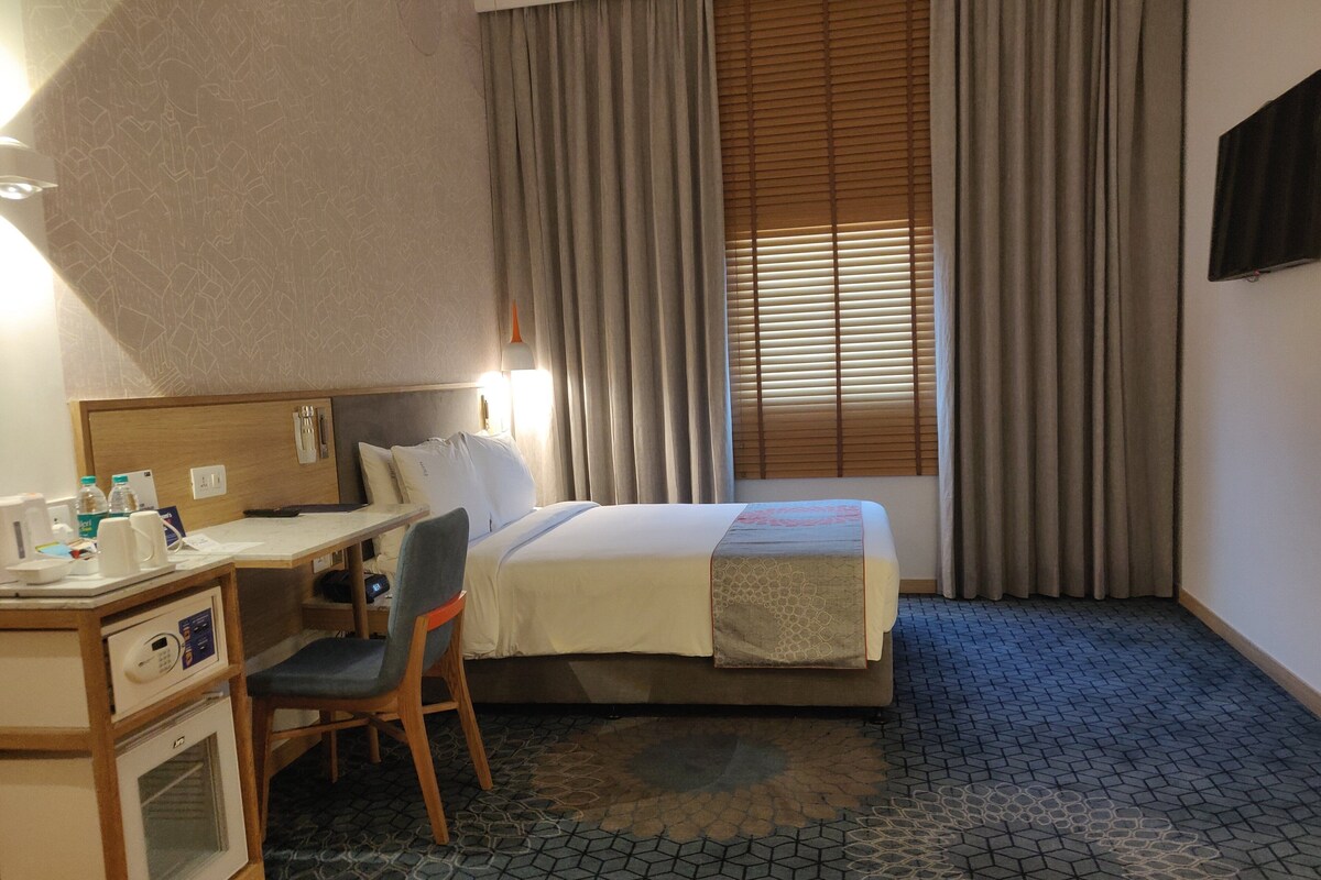 A hotel room with a bed, desk, chair, and a small kitchenette.