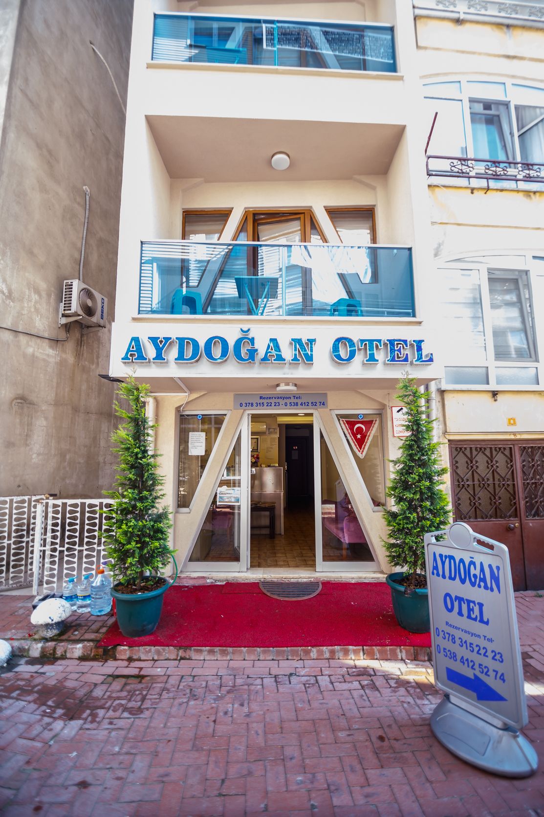 The exterior of Aydogan Otel with a red carpet entrance, two potted plants, and a signboard with contact information.