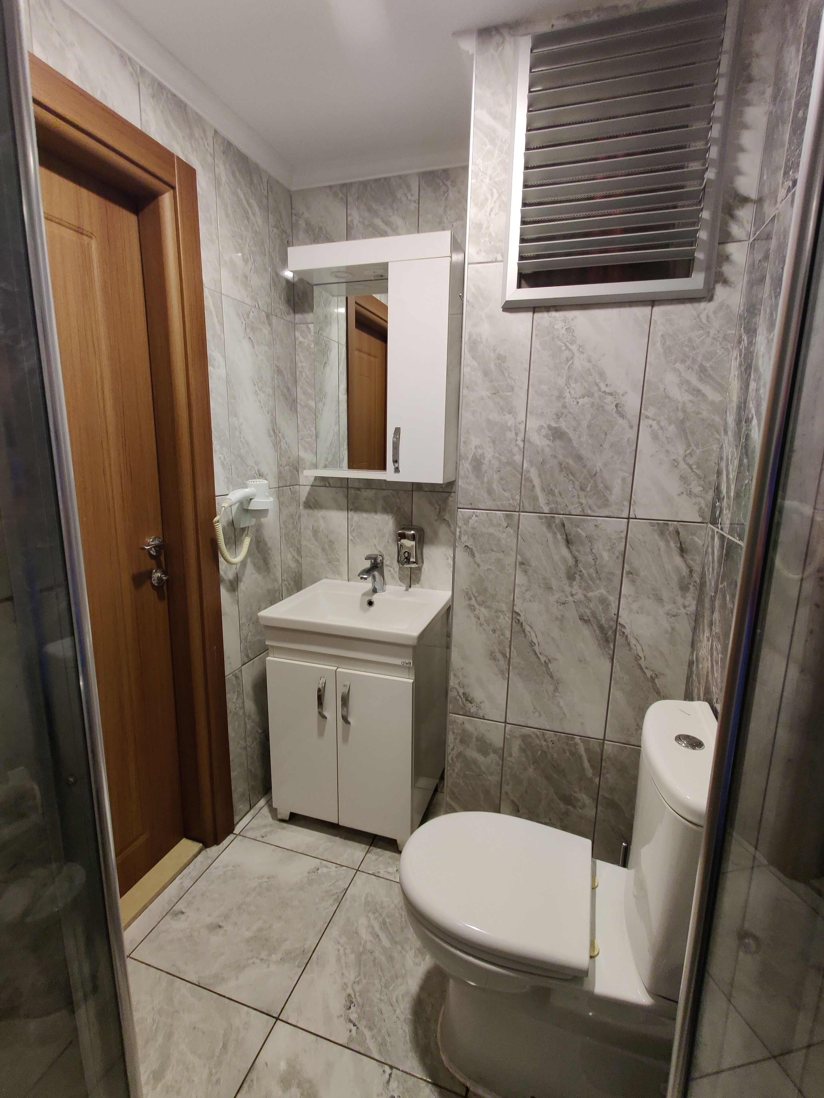 A bathroom with a wooden door, a white sink, a mirror, a toilet, and a window with blinds.