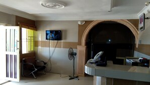 A reception area with a flat-screen TV mounted on the wall, a desk with a telephone, and a fan. - Gen-Preston Hotel & Suites (Papalanto)