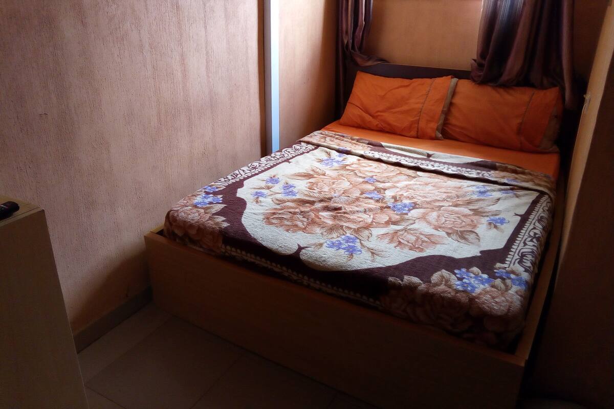 Una habitación pequeña con cama individual, colcha estampada, almohadas anaranjadas y ventana con cortinas.