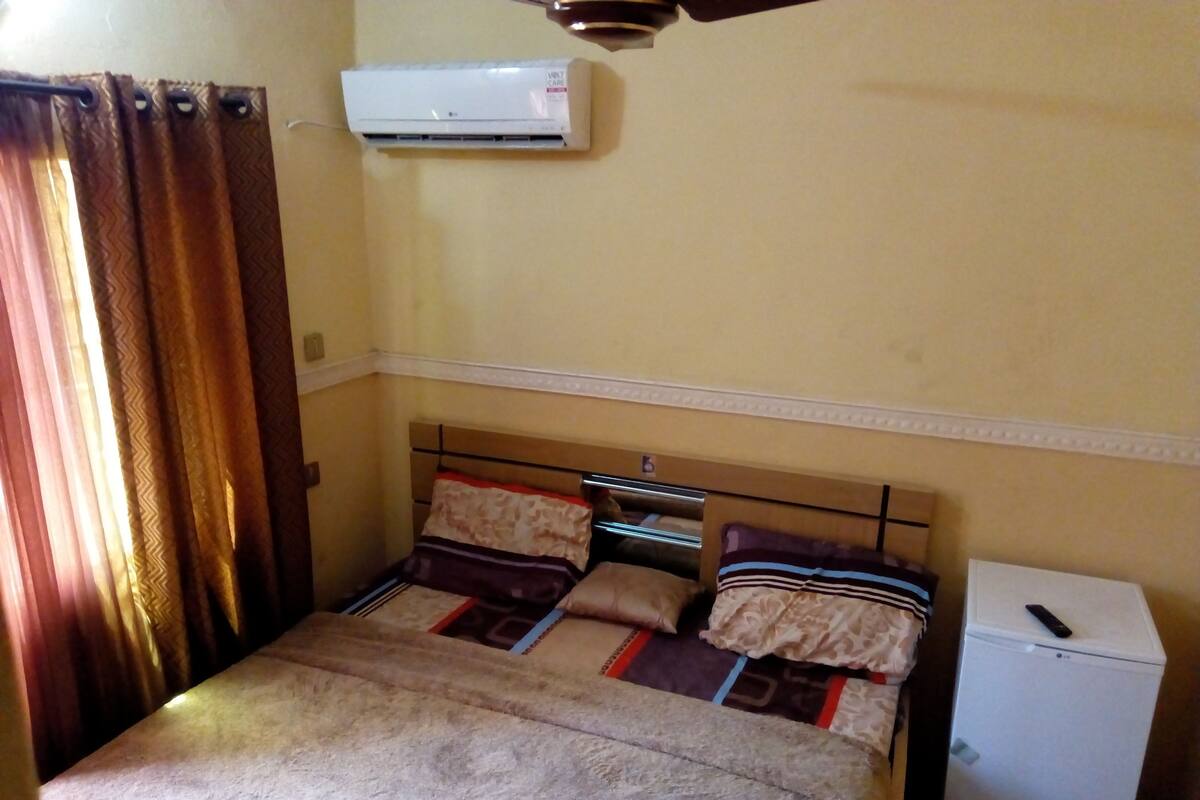 Un dormitorio con cama, un refrigerador blanco pequeño y un equipo de aire acondicionado instalado en la pared.