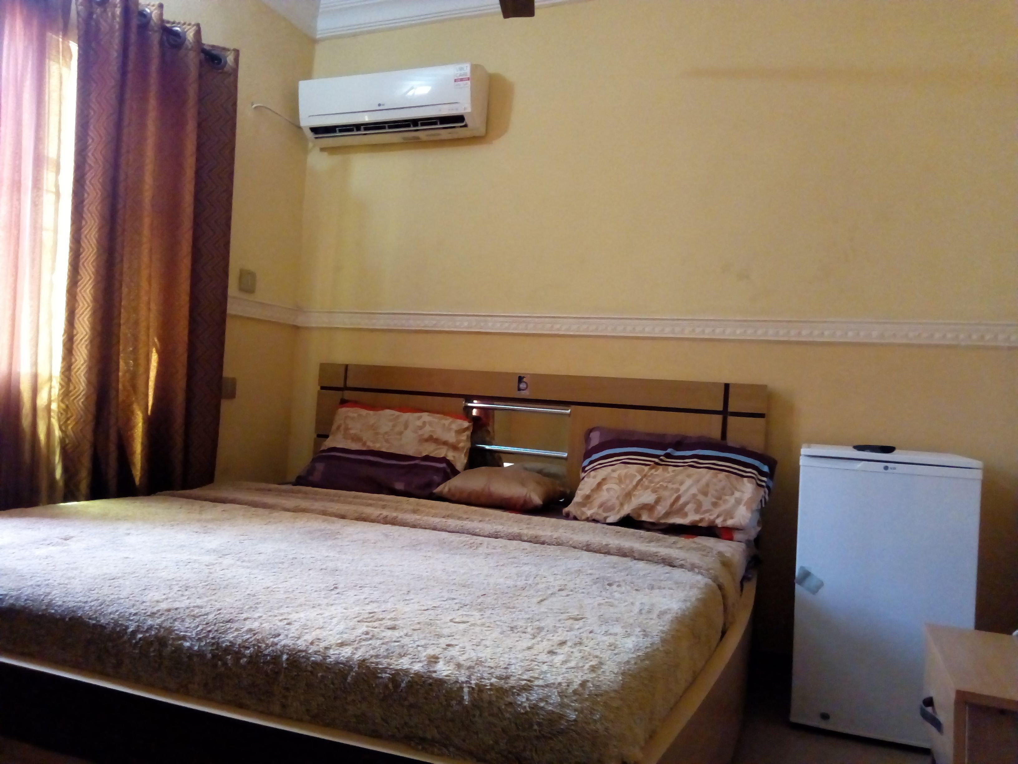 A bedroom with a bed, pillows, an air conditioner, and a small refrigerator.