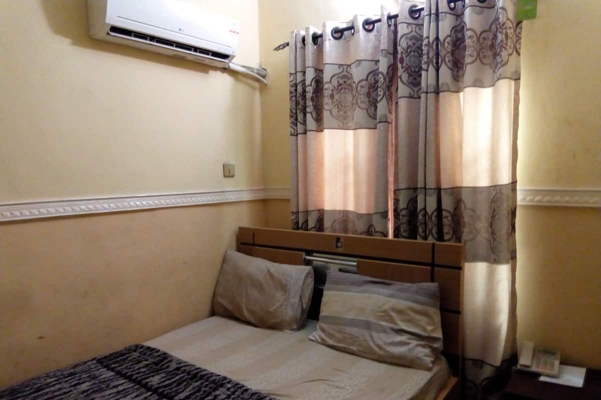 Una cama individual con cabecera de madera, una colcha estampada y dos almohadas. Un equipo de aire acondicionado está montado en la pared sobre la cama.