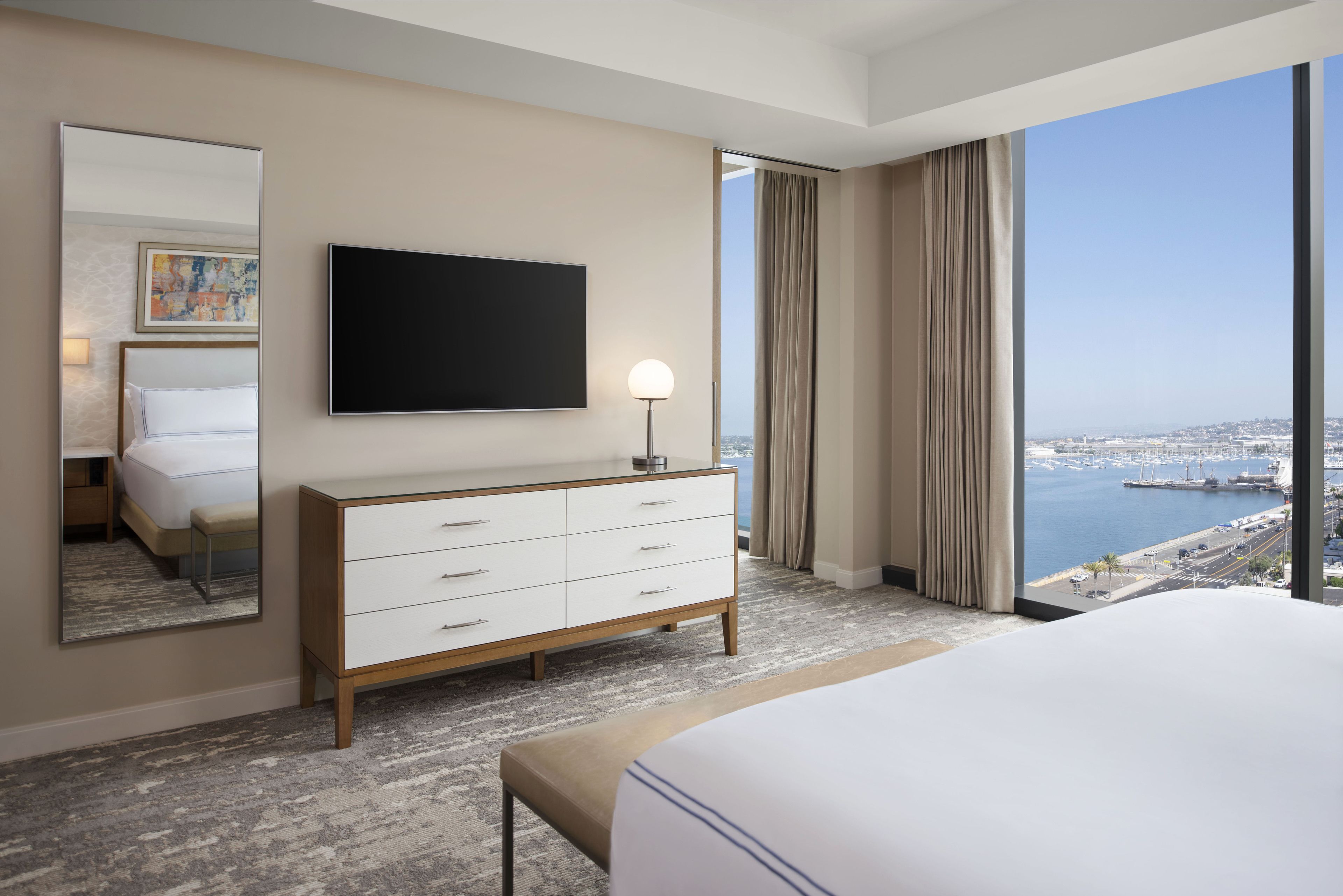 A hotel room with a large bed, a TV mounted on the wall, a dresser, and a view of the marina through large windows.