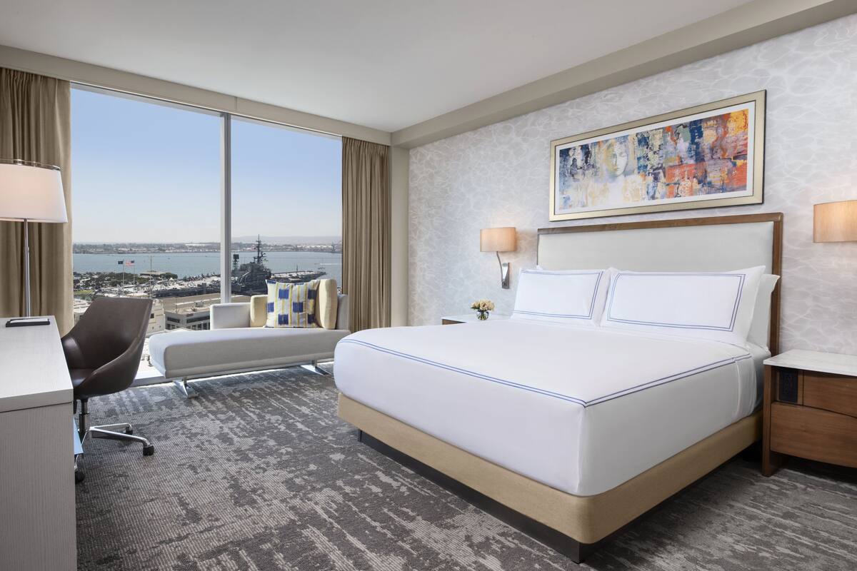 A hotel room with a large bed, a desk, a chair, and a view of a body of water and ships.