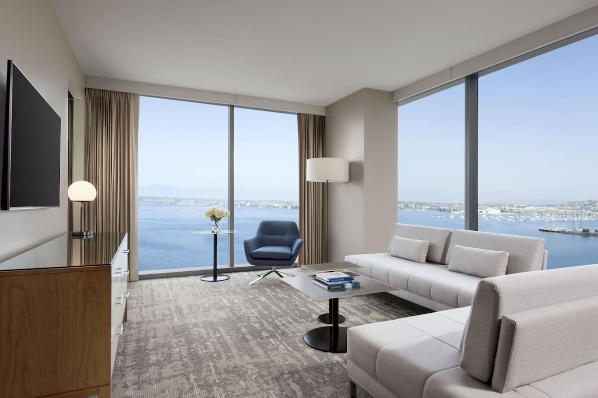 A modern hotel room with a large window, a sofa, a coffee table, a chair, and a TV.