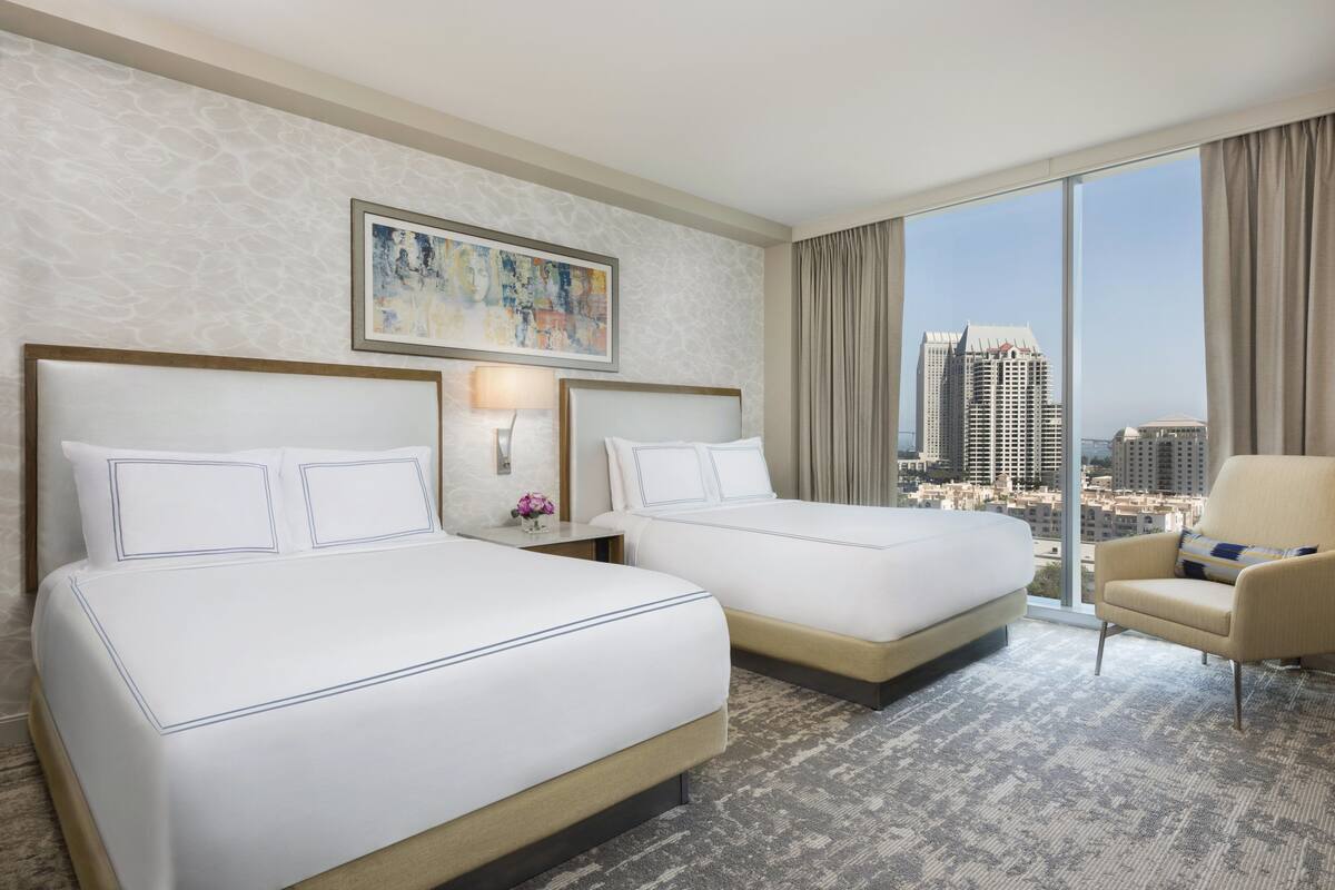 A hotel room with two beds, a large window with a city view, a painting on the wall, and a chair.
