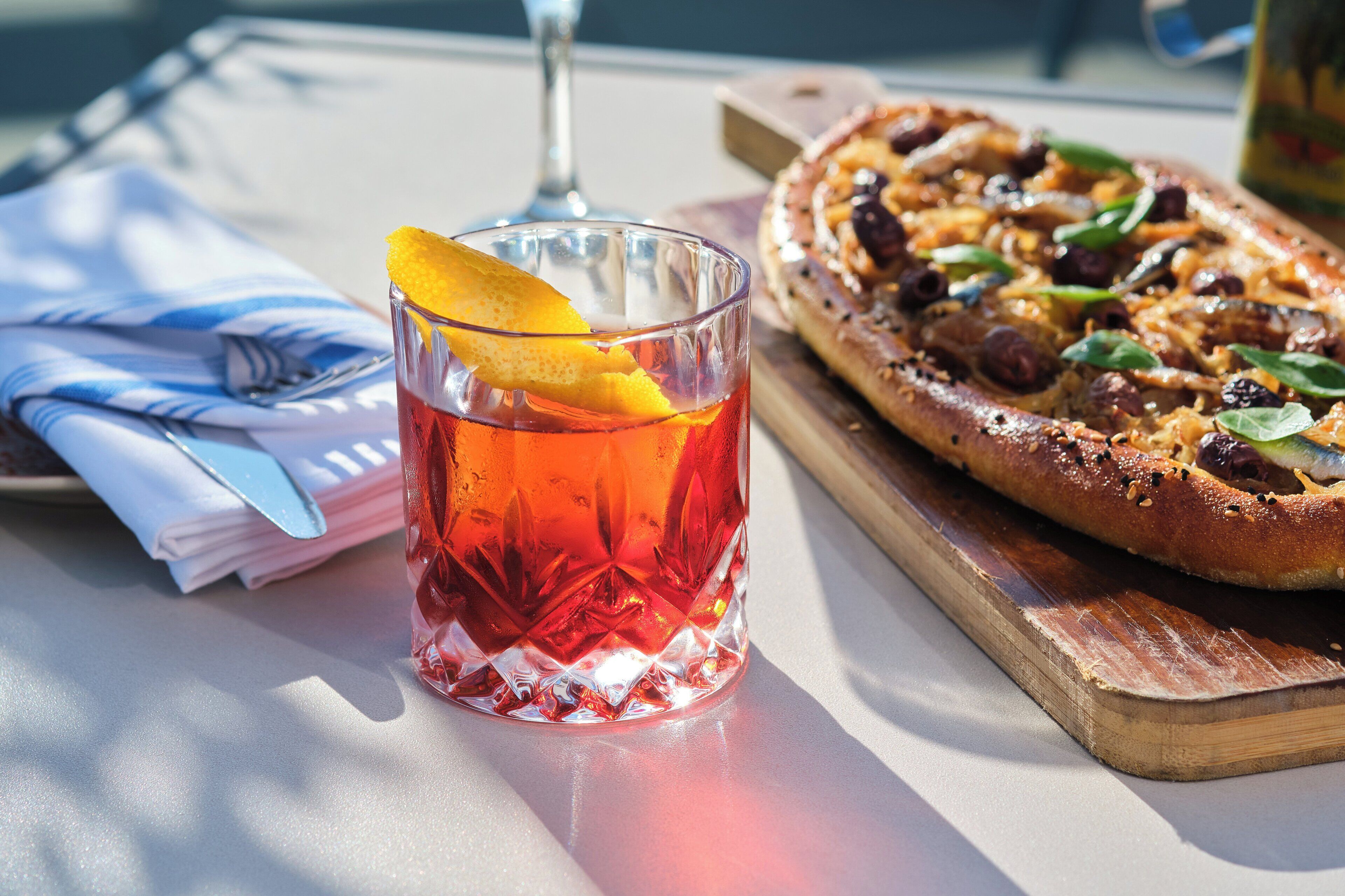 A glass of red beverage with an orange slice garnish, a pizza on a wooden board, and a napkin with a fork on a table.