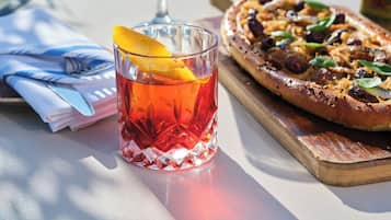 A glass of red beverage with an orange slice garnish, a pizza on a wooden board, and a napkin with a fork on a table.