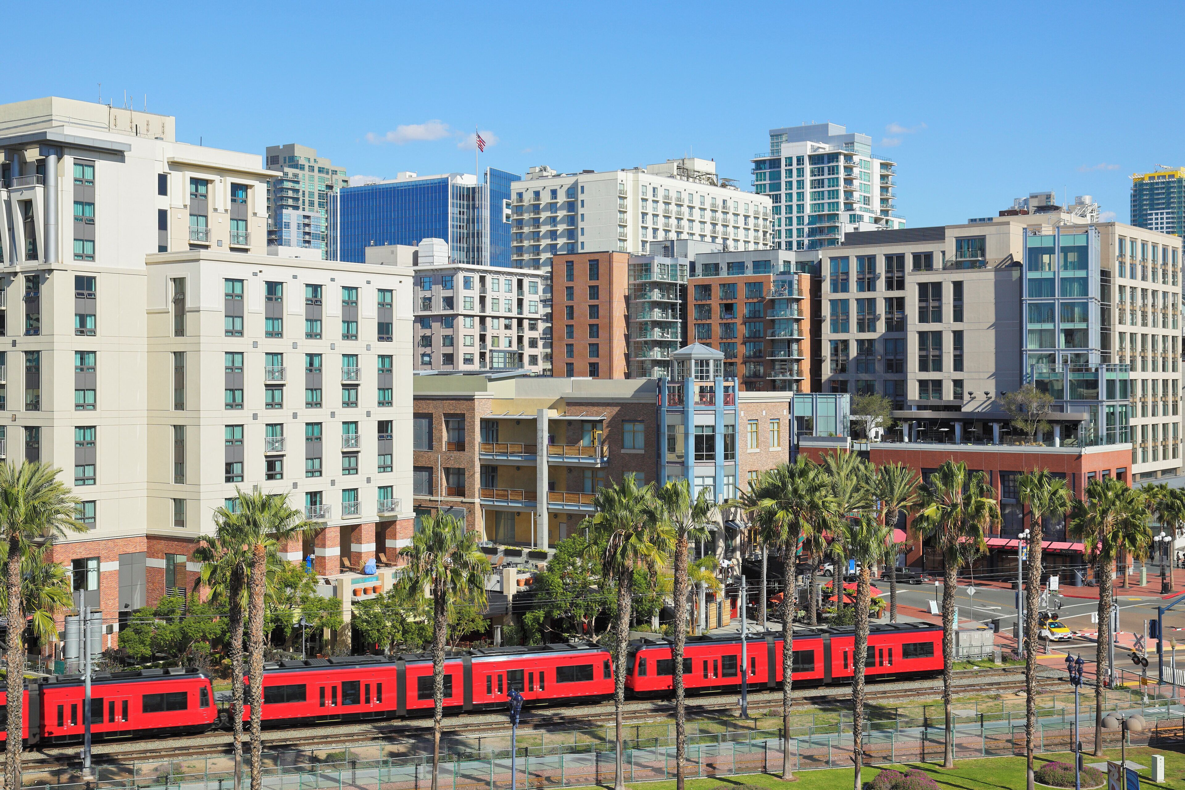 A red train passing through a cityscape with modern buildings and palm trees.