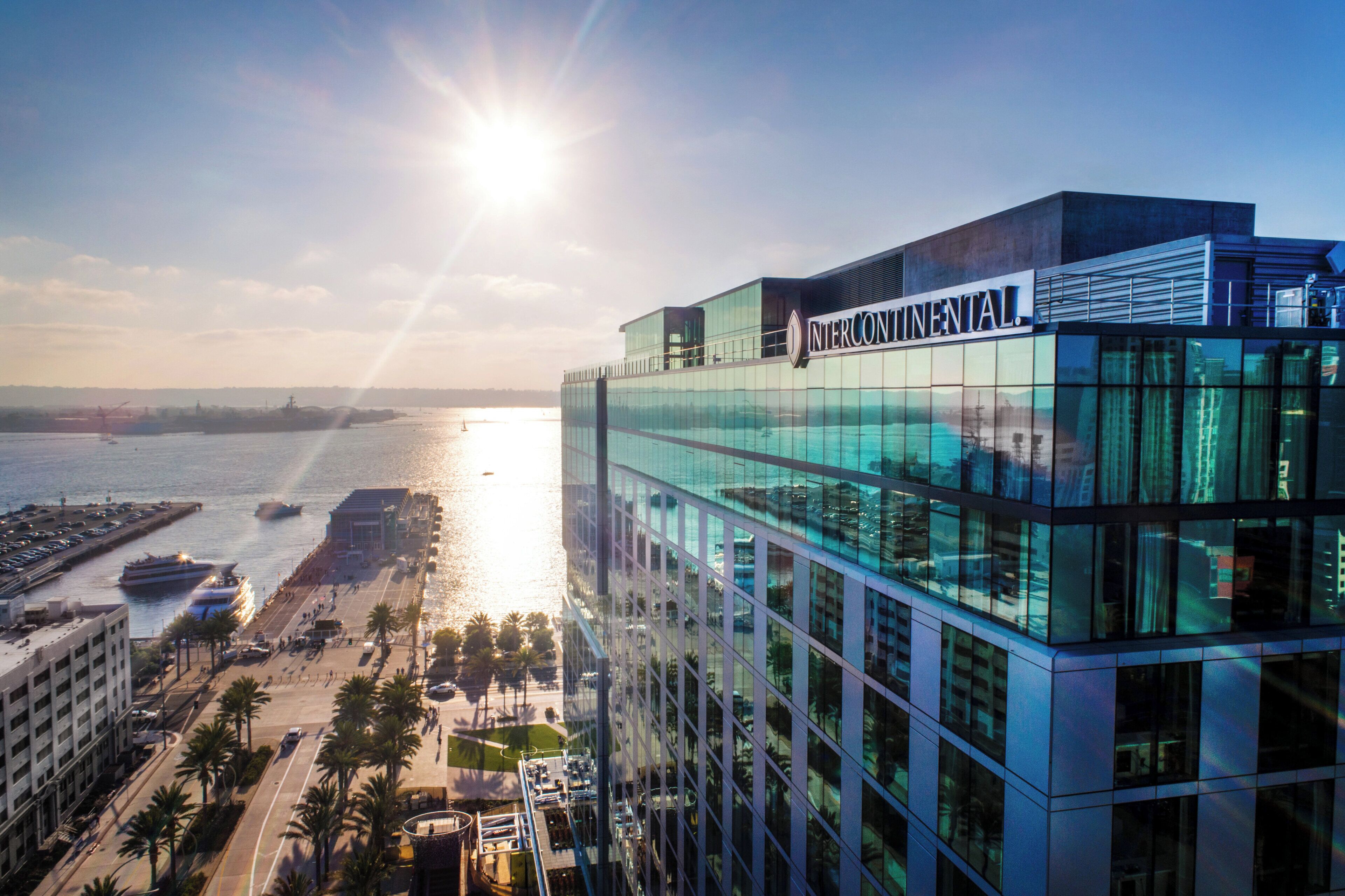A modern building with a glass facade, a clear view of the waterfront, and a parking area with cars and palm trees.