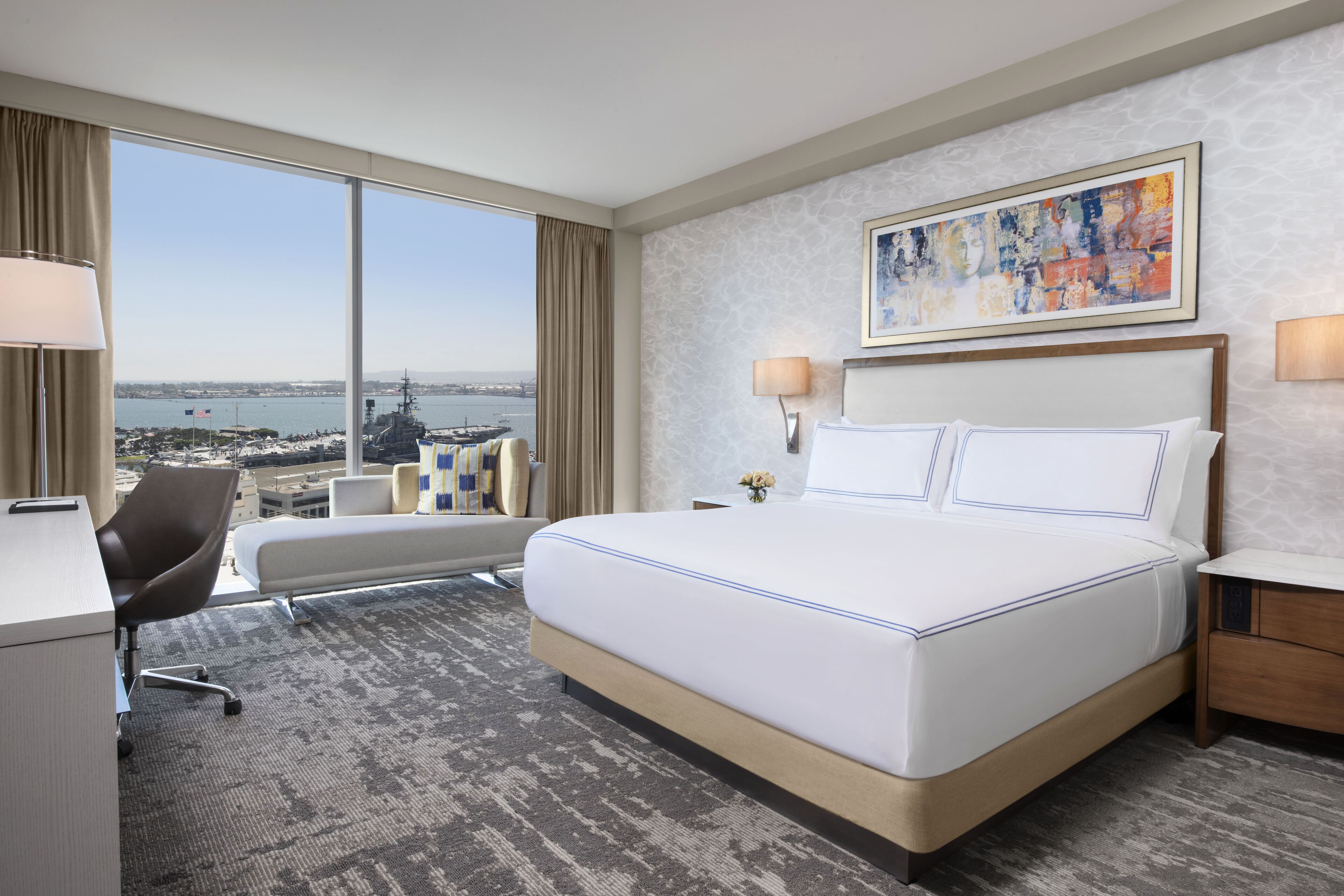 A hotel room with a large bed, a desk, a chair, and a view of a body of water and ships.