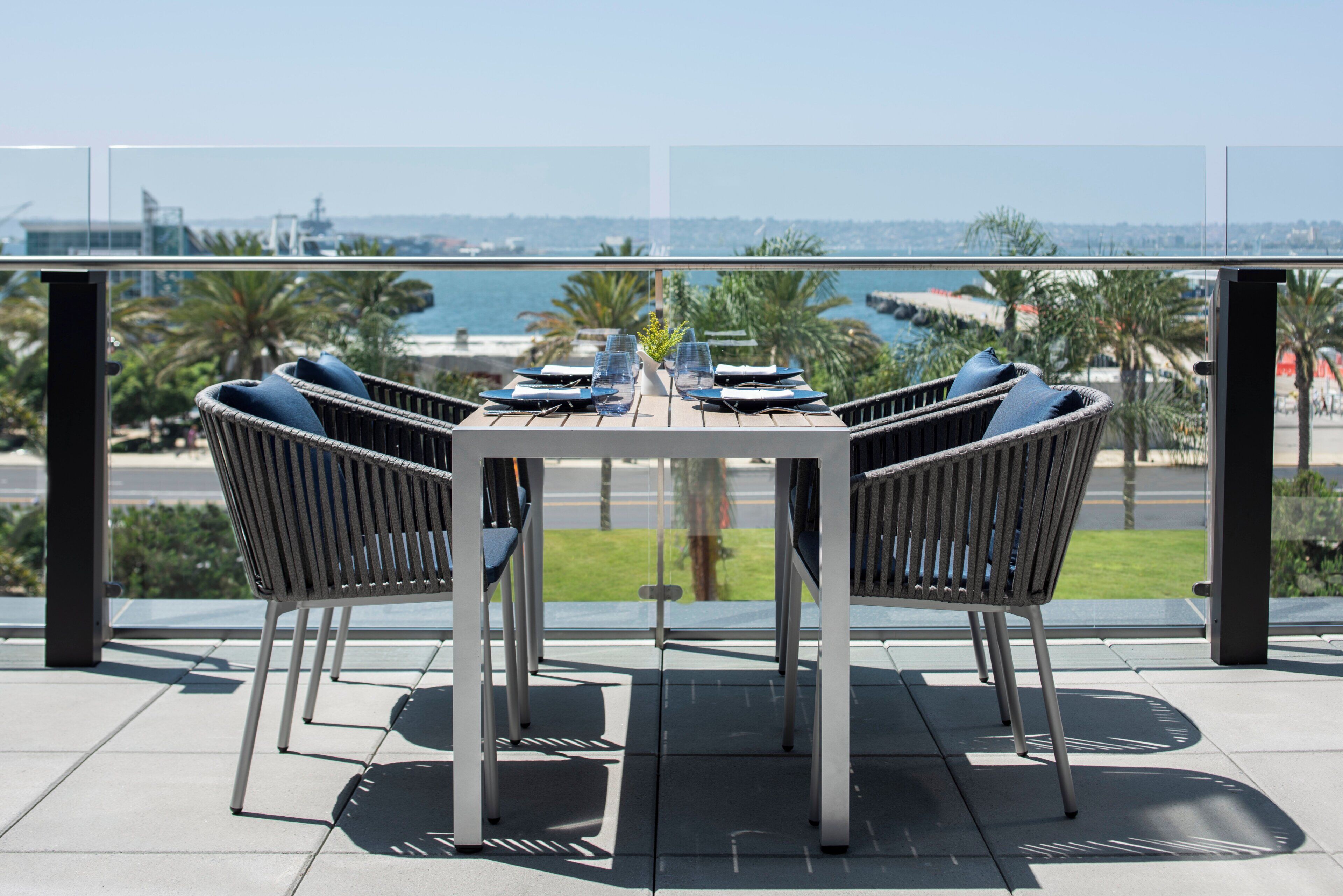 Un área de comedor al aire libre con una mesa dispuesta para cuatro personas, rodeada de sillas de mimbre con cojines y vistas a la costa.