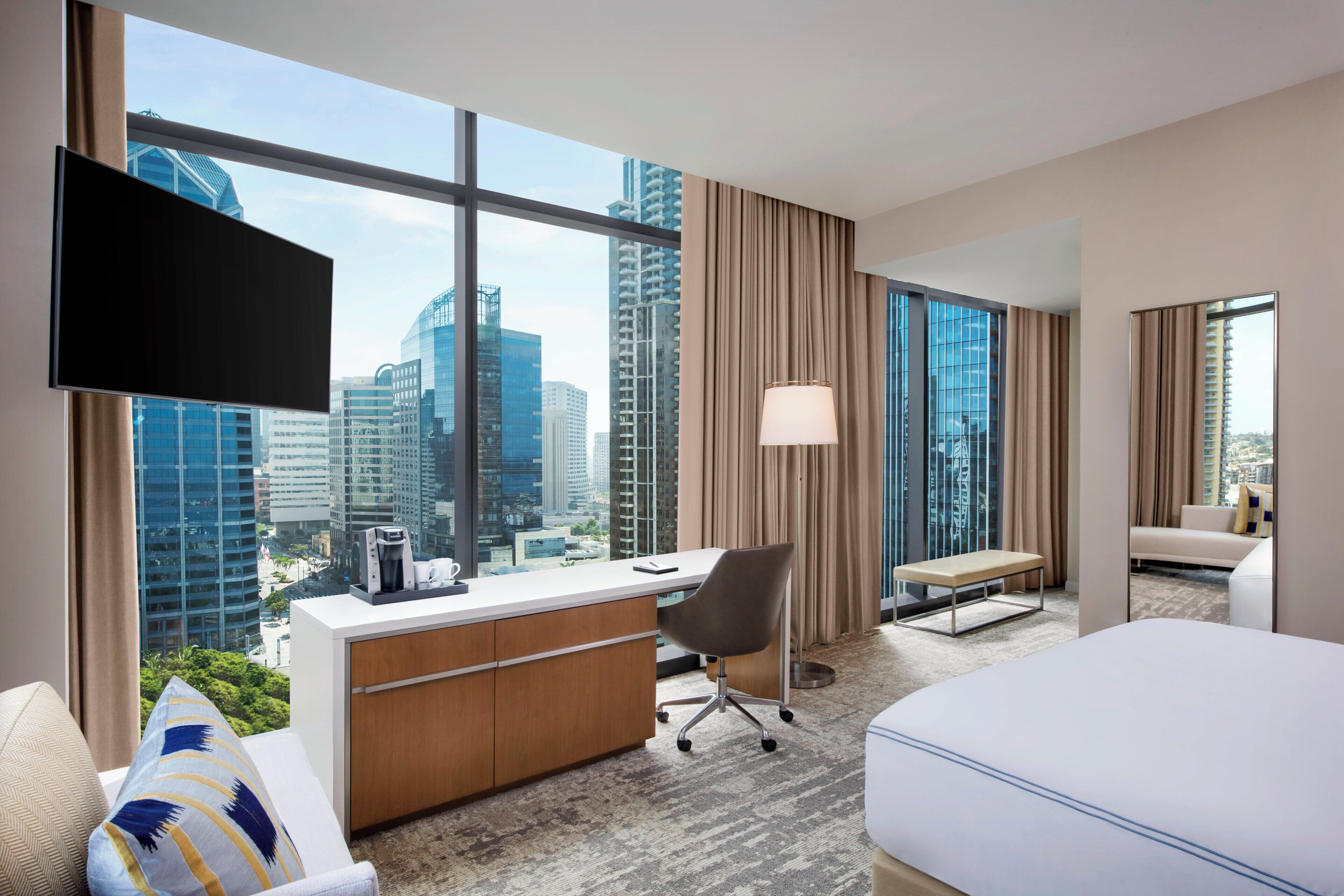 Una habitación de hotel con un ventanal grande, un escritorio, una silla, una cama y un televisor.