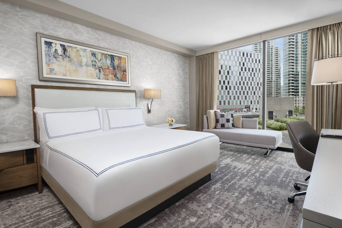A hotel room with a large bed, a desk, a chair, a sofa, and a view of tall buildings.