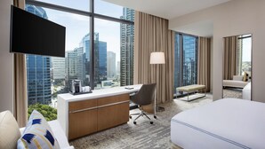 A hotel room with a large window, a desk, a chair, a bed, and a TV.