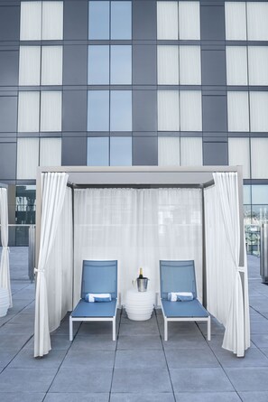Una terraza en la azotea con dos sillones, una mesita redonda con una botella y vistas a un edificio moderno con grandes ventanales.