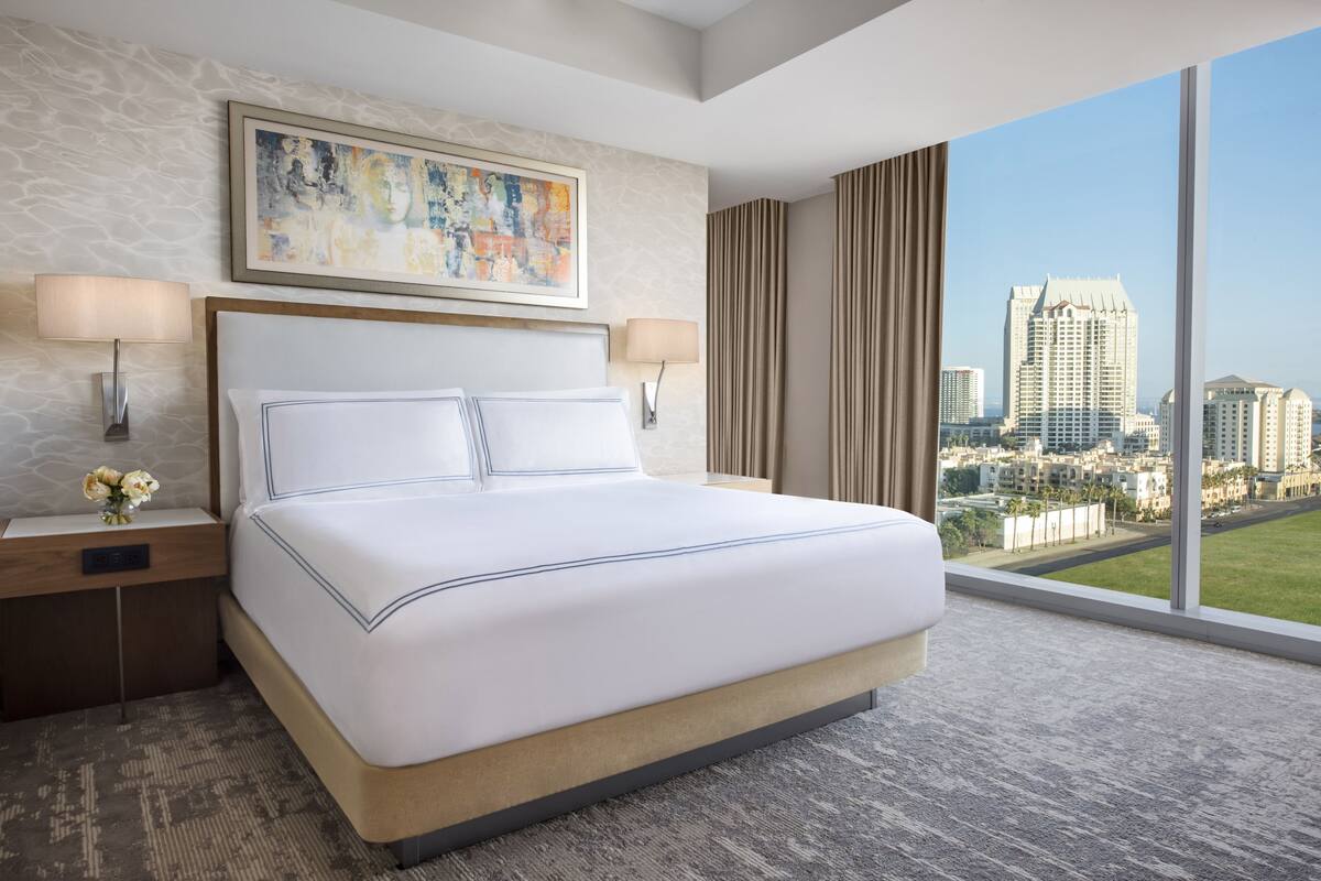 A hotel room with a large bed, two bedside lamps, a nightstand with a vase of flowers, a painting above the bed, and a city view through large windows.