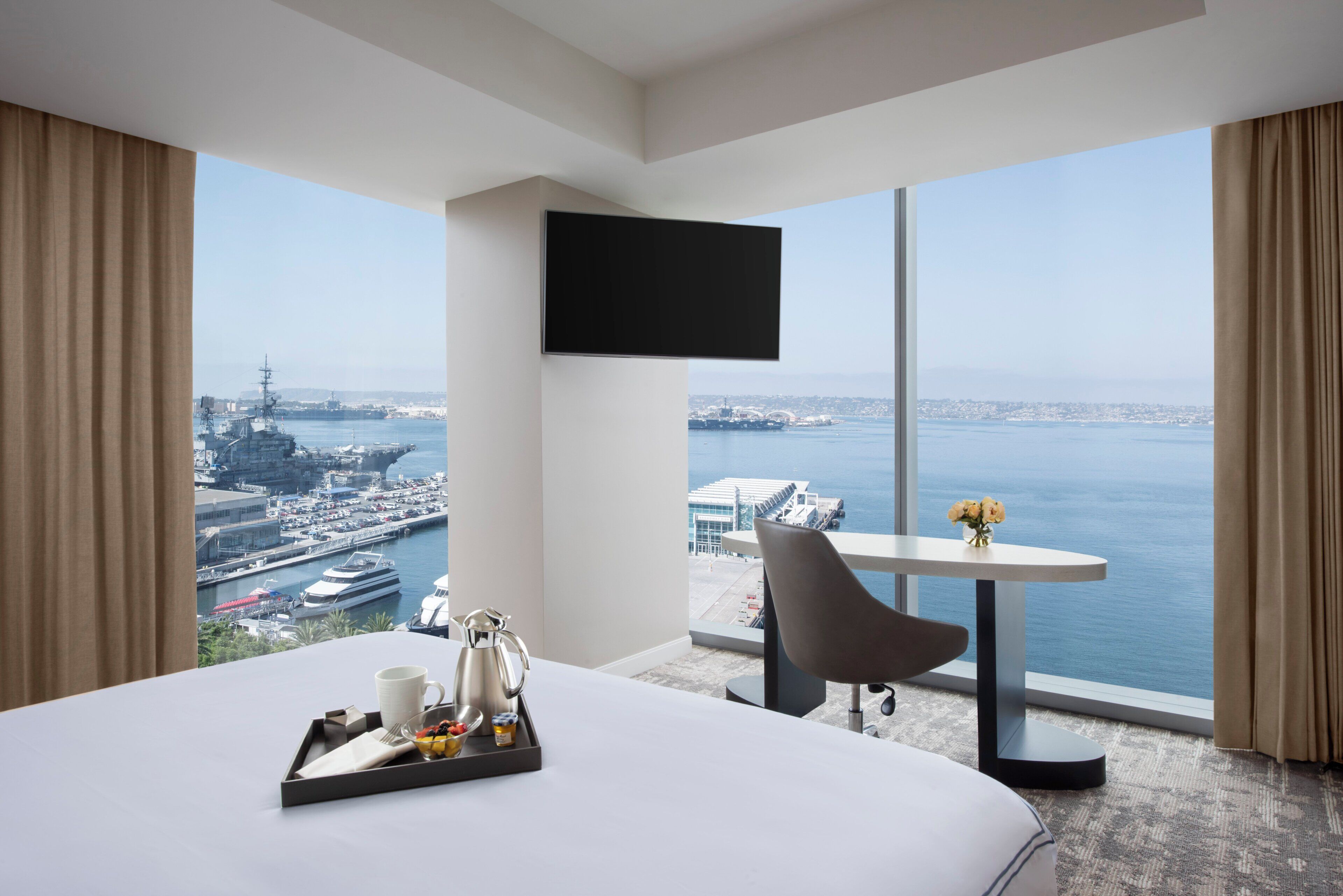 Habitación de hotel con una cama grande, una mesa de comedor con una bandeja de desayuno, un televisor montado en la pared y vistas al mar y al paisaje urbano.