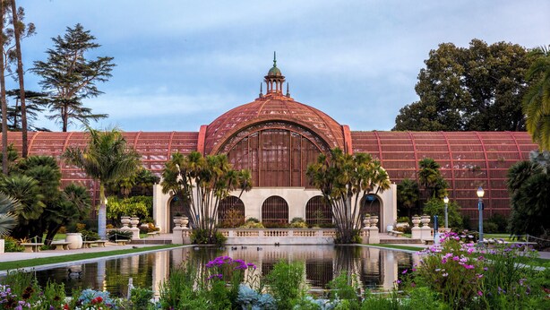 A large building with a central dome and arched windows, surrounded by a reflective pond and colorful flowers.