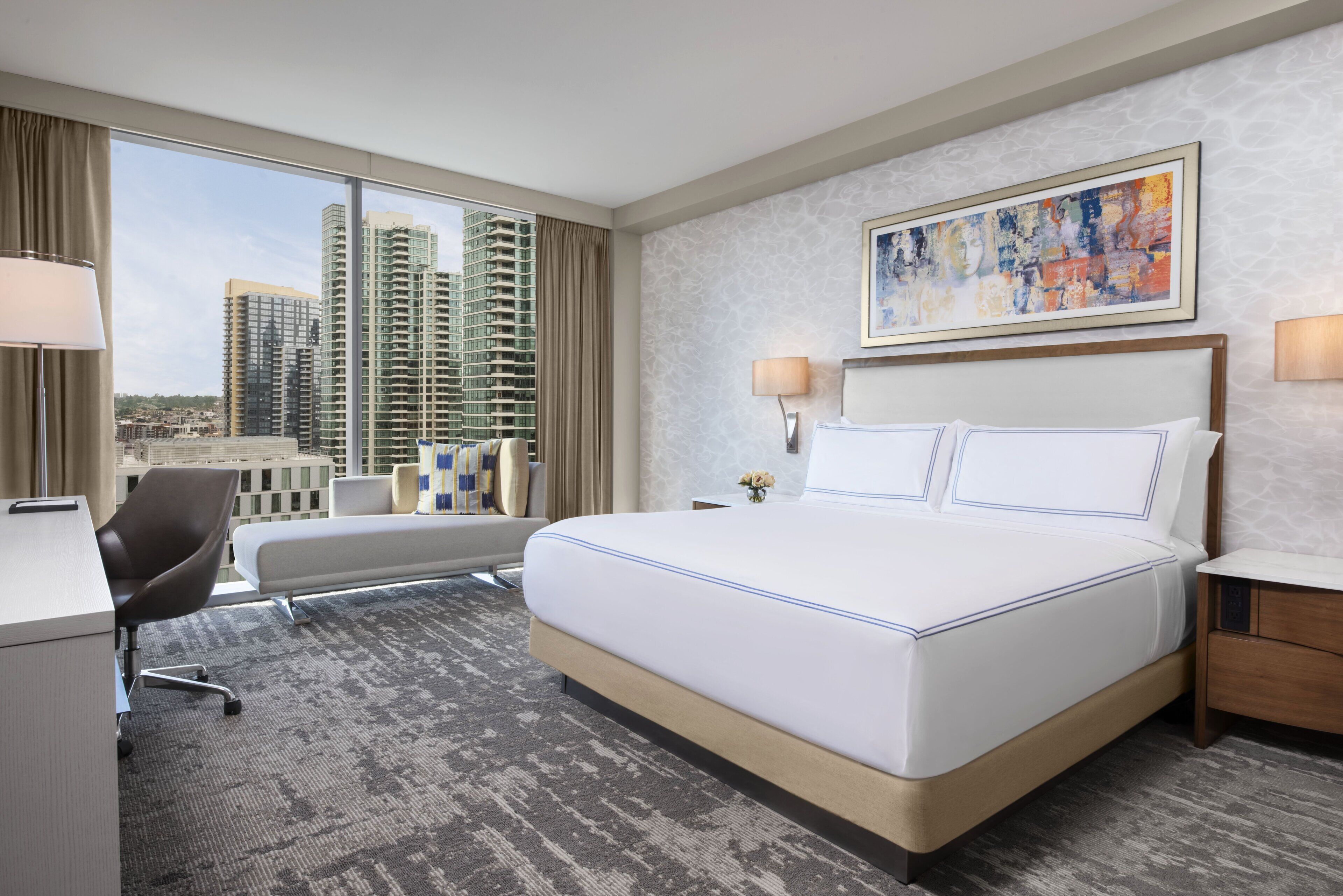 A hotel room with a large bed, a desk, a chair, and a view of tall buildings.