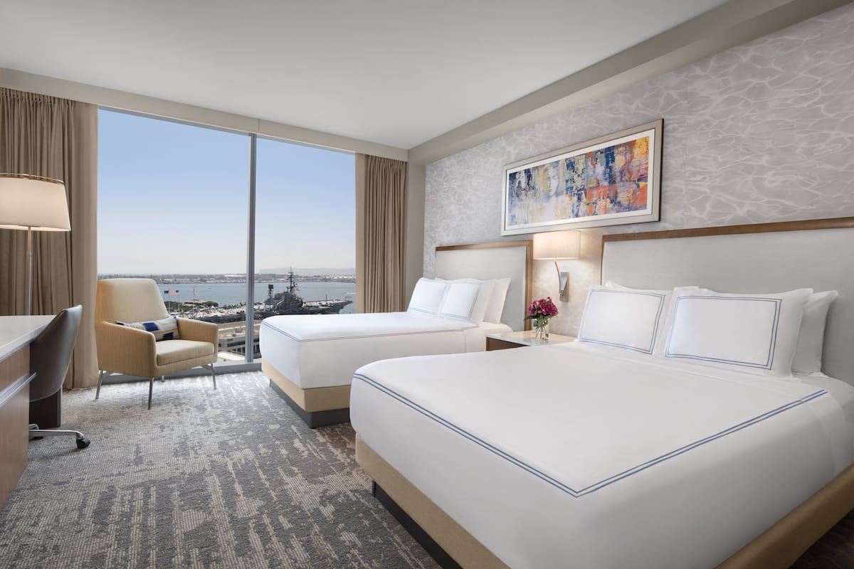 A hotel room with two beds, a desk, a chair, and a large window offering a city view.
