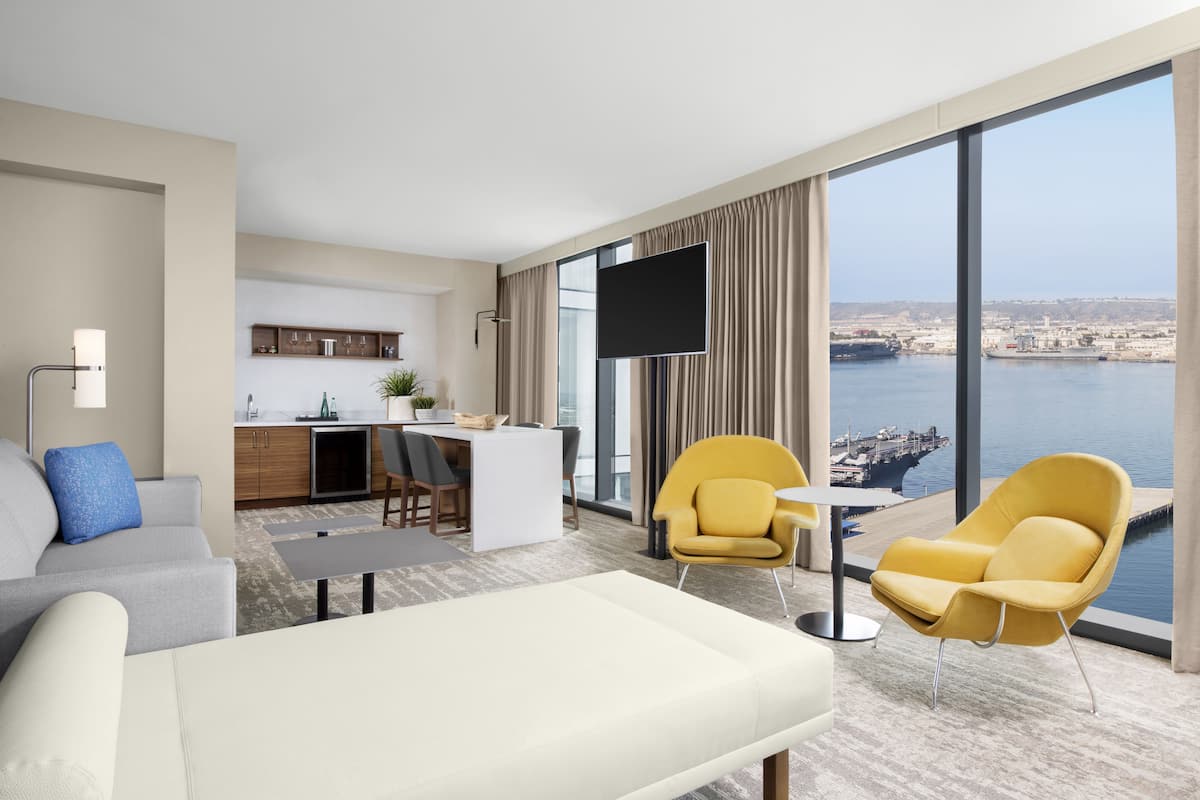 A modern hotel room with a large window offering a city view, a sofa, a yellow armchair, a small dining table, and a flat-screen TV.