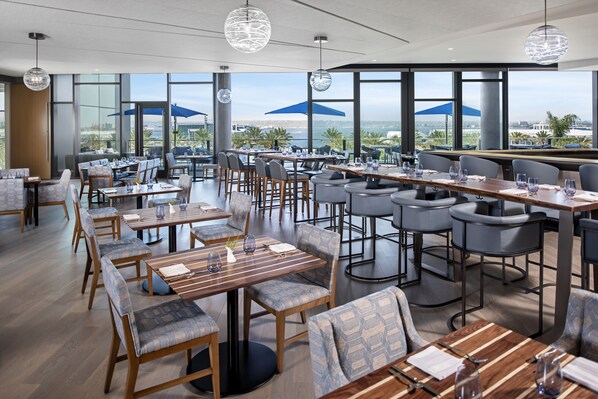 A modern restaurant with multiple dining areas, large windows offering ocean views, and contemporary lighting fixtures.