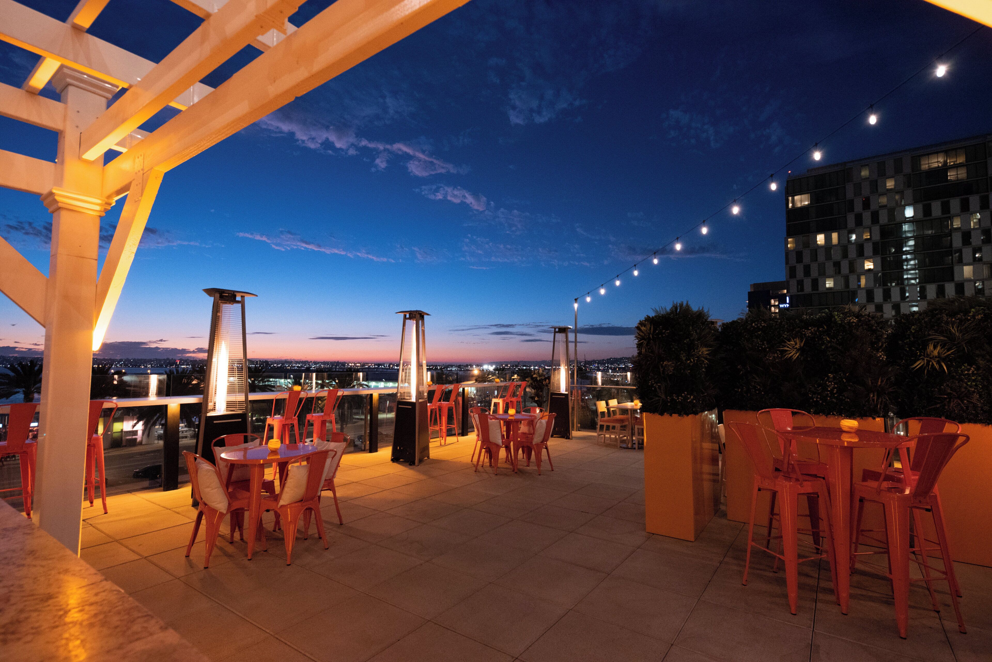 An outdoor terrace with tables and chairs, a pergola structure, and a view of the cityscape at dusk.