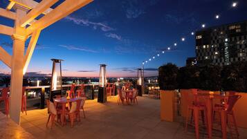 An outdoor terrace with tables and chairs, a pergola structure, and a view of the cityscape at dusk.
