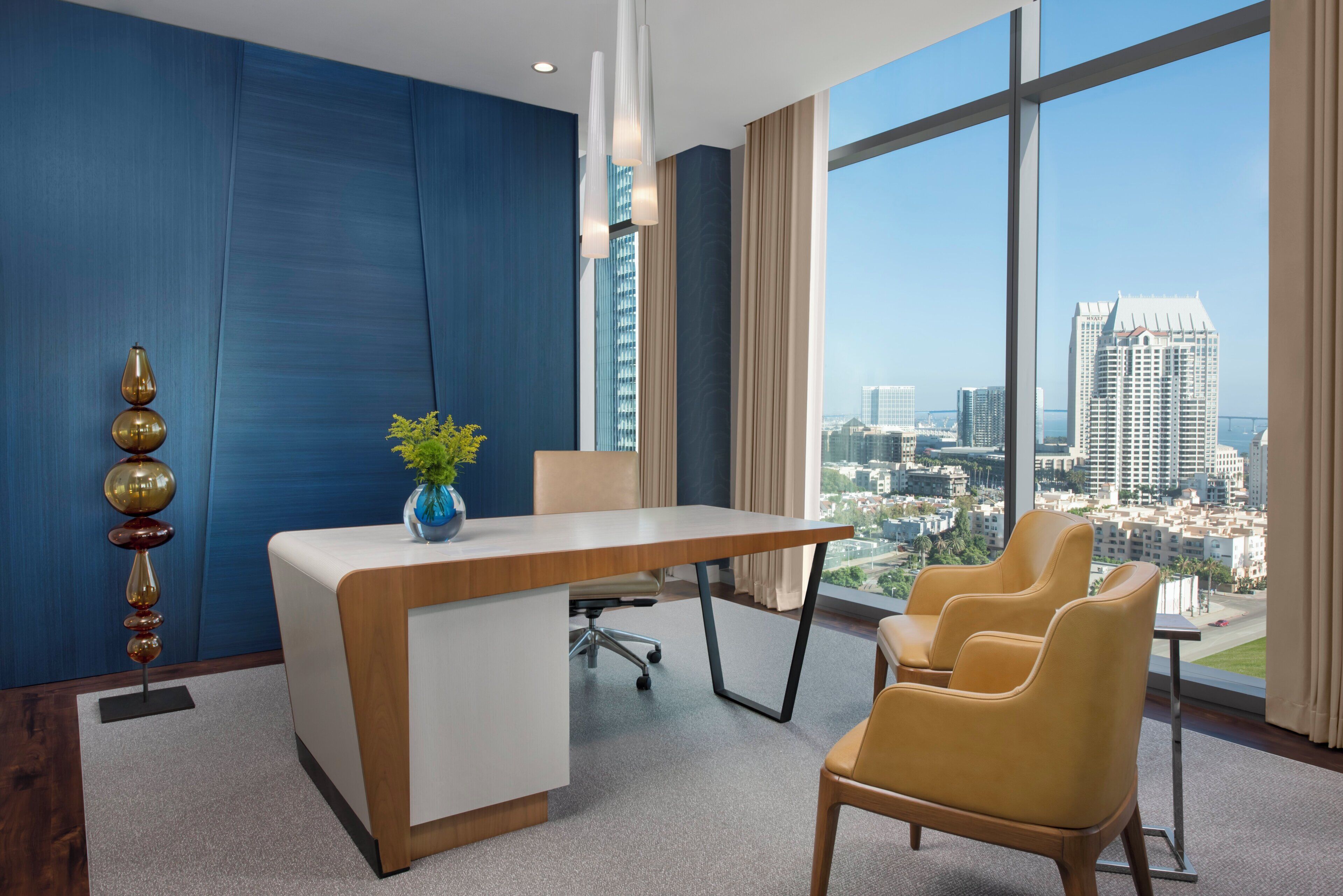 A modern office space with a large desk, chairs, a vase with flowers, and a city view through large windows.