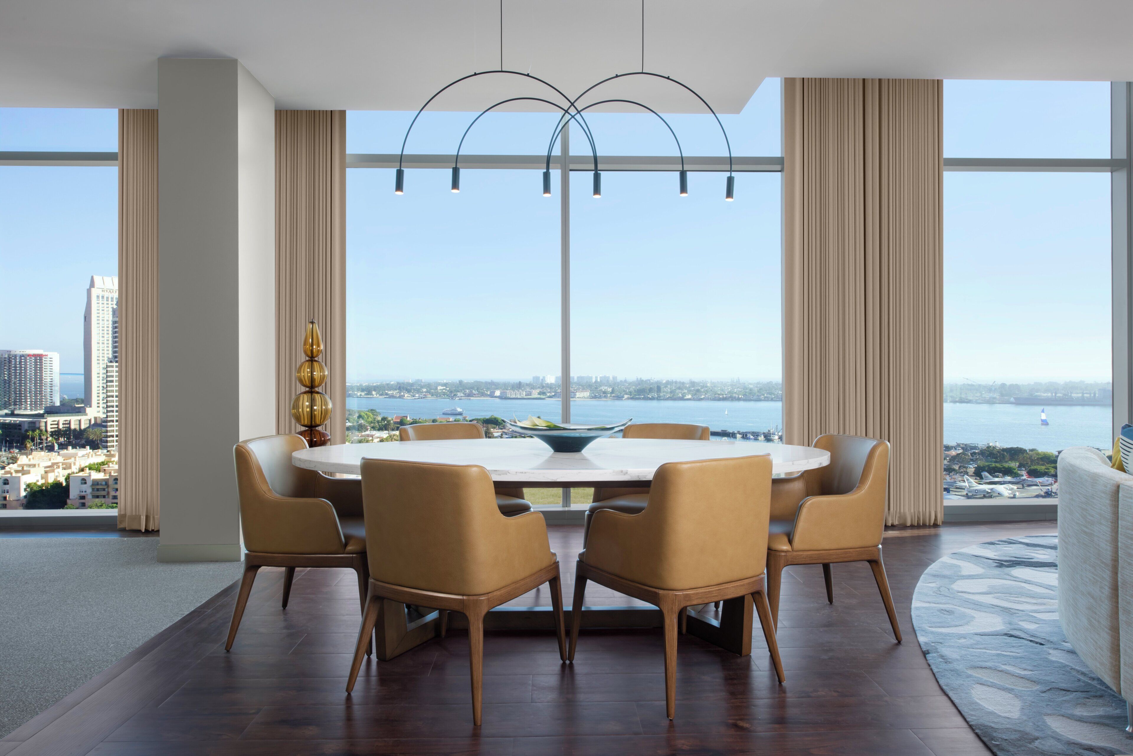 A modern dining room with a round table, six chairs, and a view of a cityscape and water.