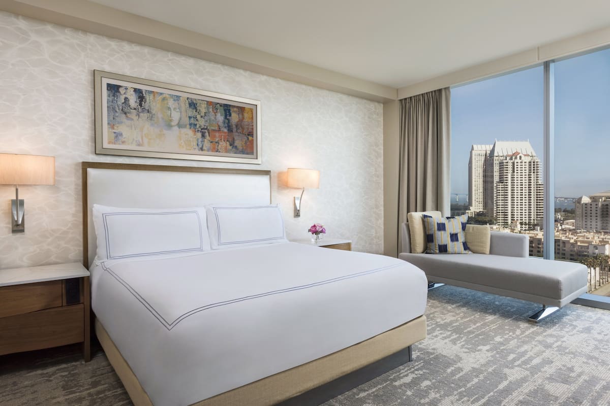 A hotel room with a large bed, two bedside tables with lamps, a sofa, a large window with a city view, and a painting on the wall.