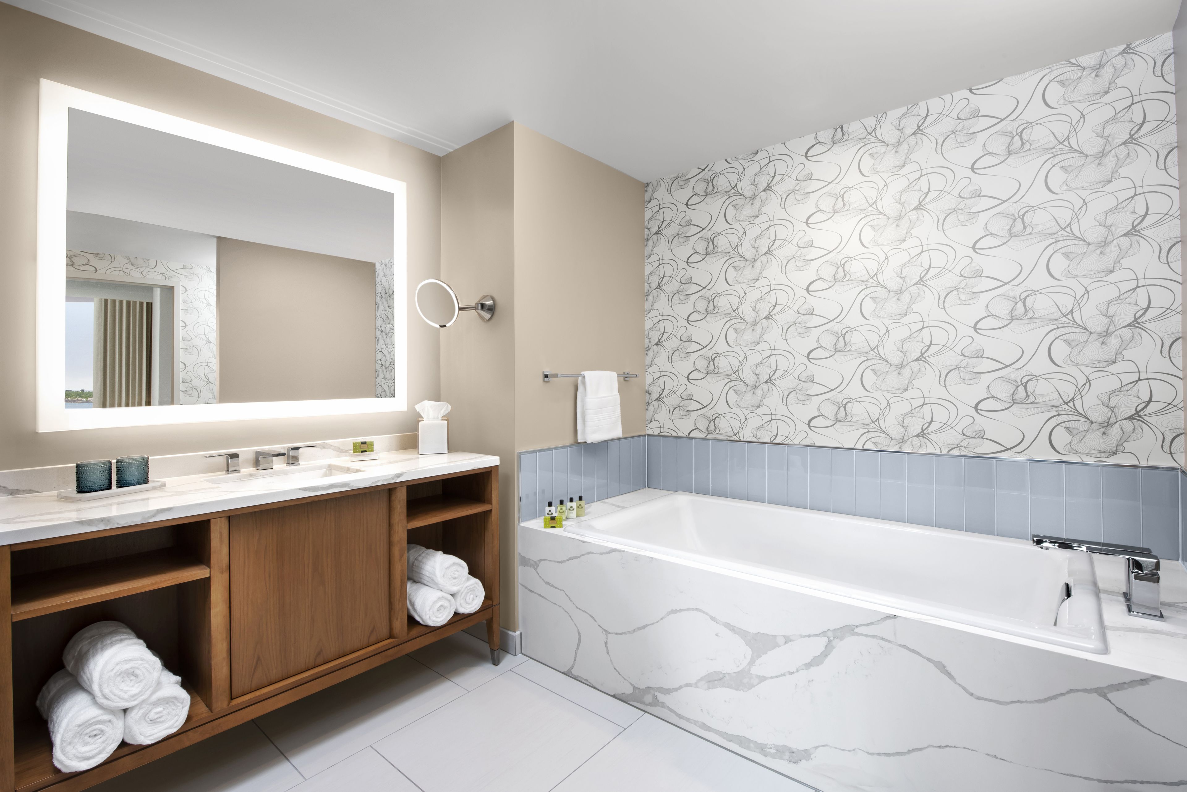 A modern bathroom with a large mirror, marble bathtub, wooden vanity, and patterned wallpaper.