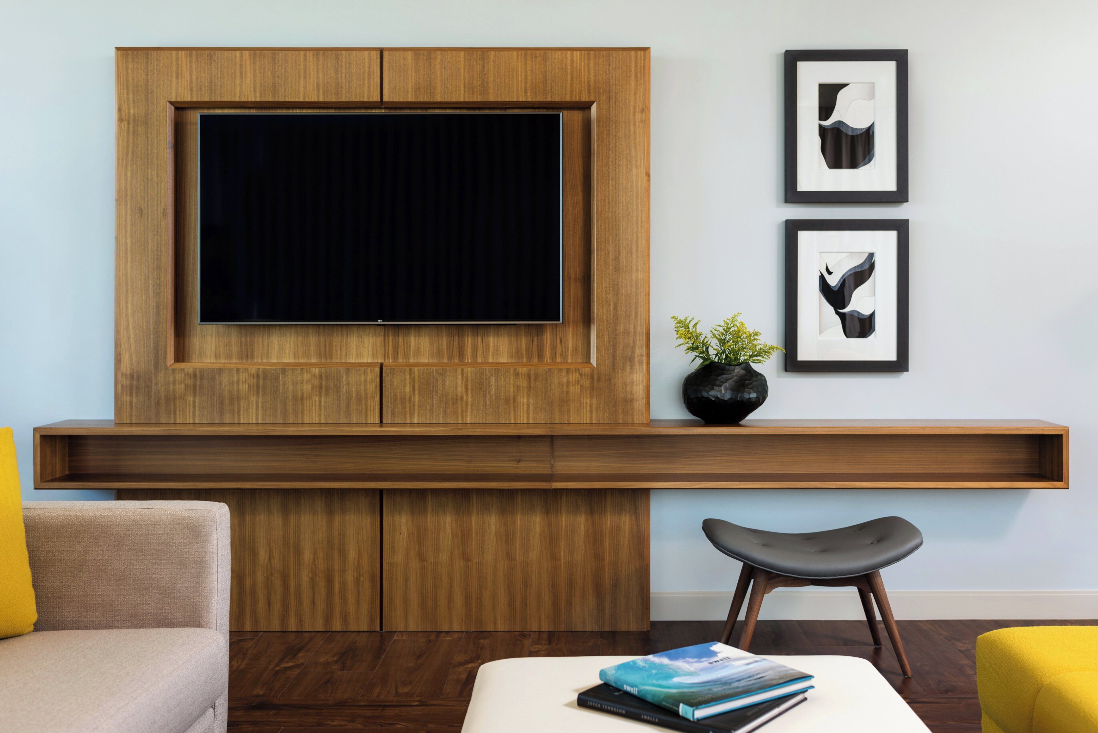 A modern living room with a large flat-screen TV mounted on a wooden panel, a small black vase with yellow flowers, and two framed abstract art pieces on the wall.