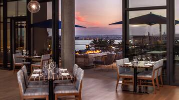Un espacio exterior con zona de comedor, mesas, sillas, un elemento decorativo de fuego y vistas al agua y al paisaje urbano.
