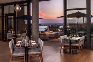 Un espacio exterior con zona de comedor, mesas, sillas, un elemento decorativo de fuego y vistas al agua y al paisaje urbano.