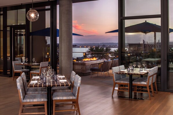 An outdoor dining area with tables and chairs, a fire feature, and a view of the water and cityscape. - InterContinental San Diego by IHG (San Diego)