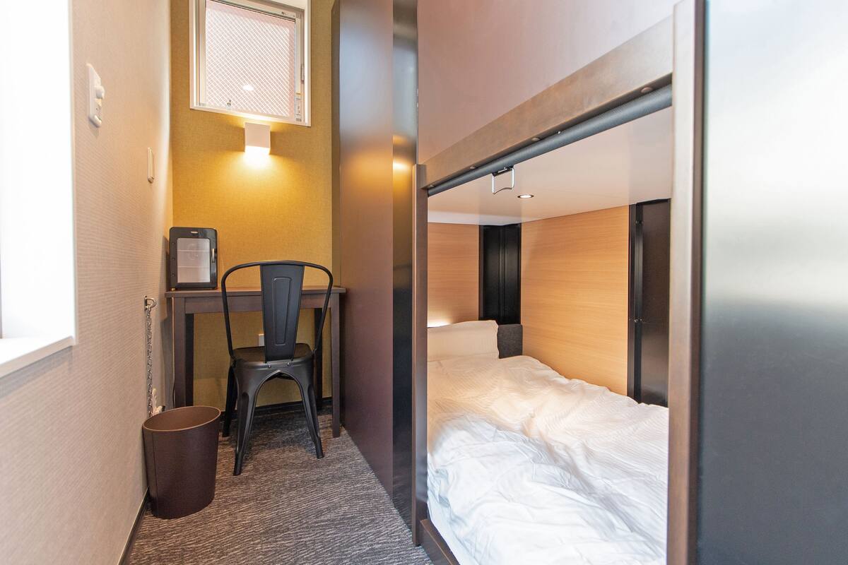 A compact hotel room with a bunk bed, a desk with a chair, and a small trash bin.