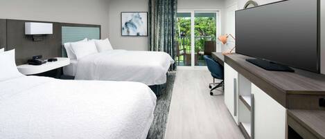 A modern hotel room with two beds, a flat-screen TV, a desk with a lamp, and a view of greenery through the window.