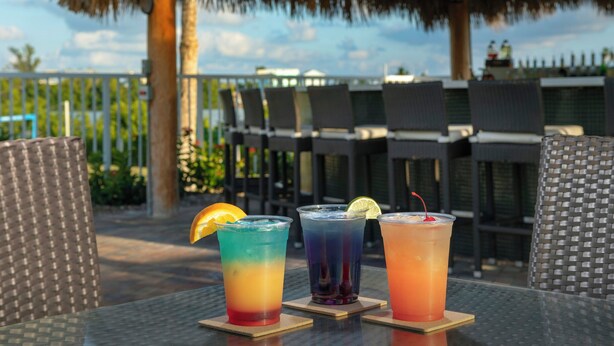 Three colorful drinks on a table with chairs and a thatched roof in the background. - Hampton Inn Marathon - Florida Keys (Marathon)