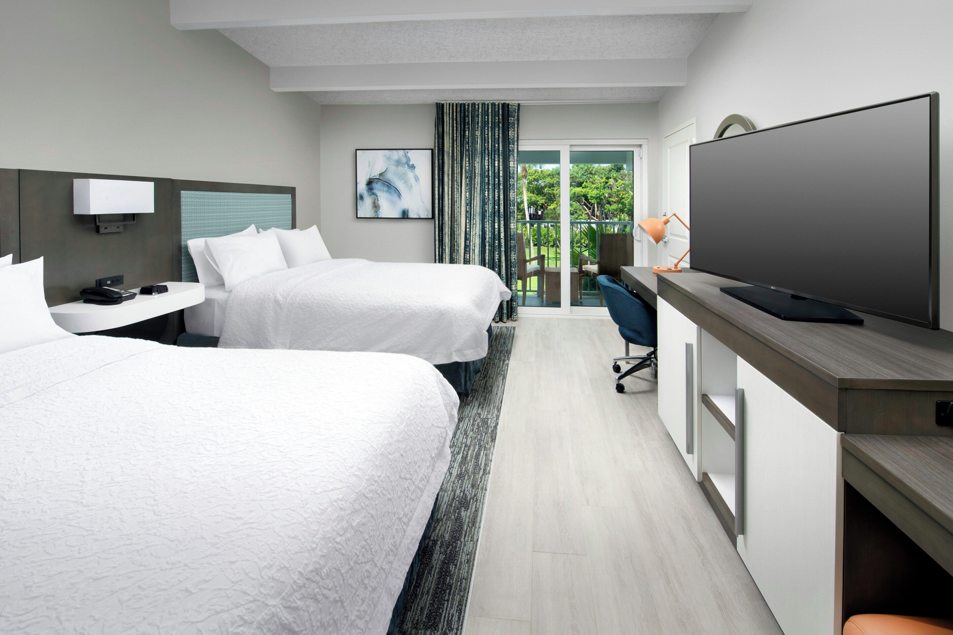 A hotel room with two beds, a TV, a desk, and a balcony with a view of greenery.