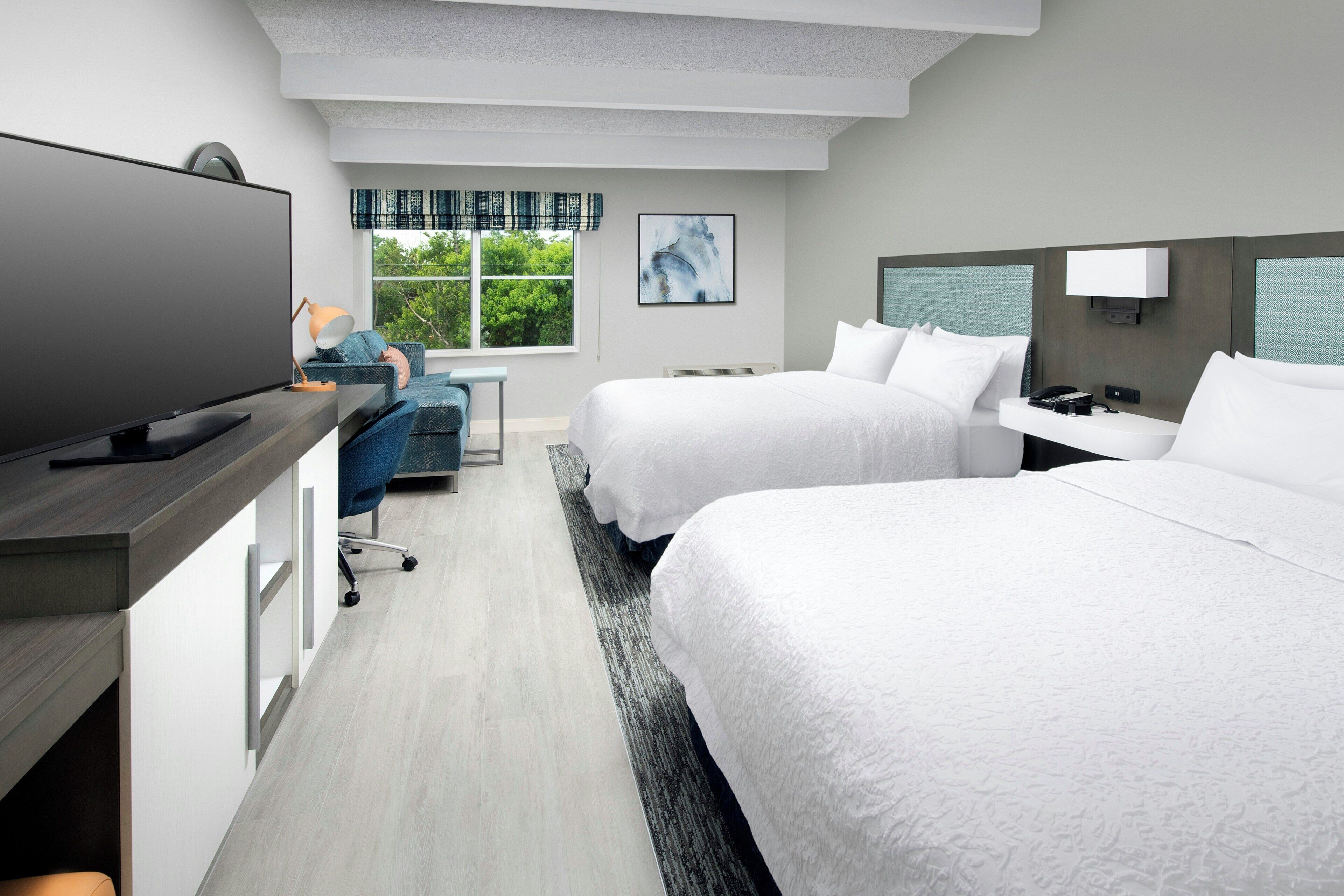 A hotel room with a flat-screen TV, two beds, a desk with a chair, and a window with a view of greenery.