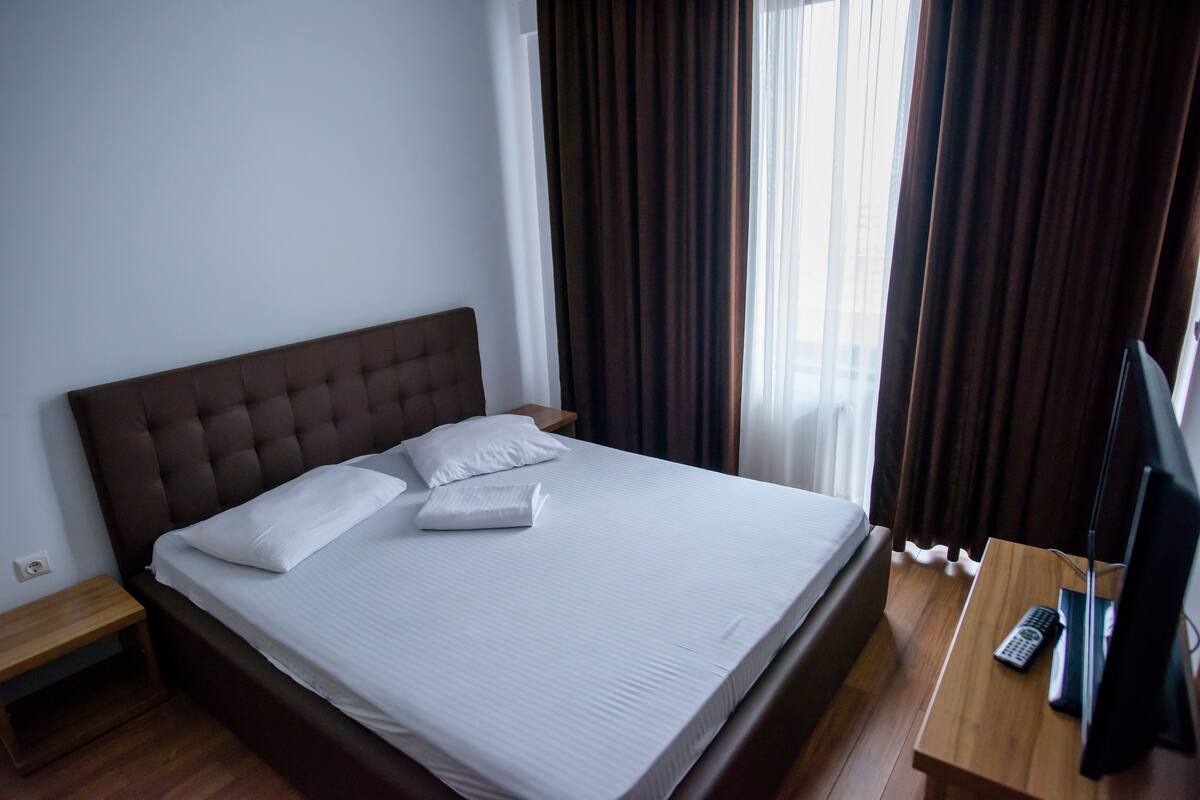 Una cama bien hecha con sábanas y almohadas blancas, una mesita de noche de madera, un televisor sobre una mesa de madera y una ventana con cortinas.